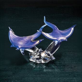Blue glass manta ray sculpture with silver detailing, layered translucent wings, and crystalline coral base against a dark textured backdrop.