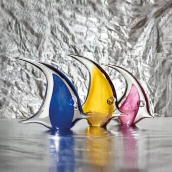 Glass fish sculptures—blue, yellow, pink—with curved fins displayed on a reflective surface against crinkled silver foil backdrop.