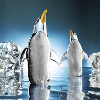 Glass penguin sculptures by Duccio Di Segna on blue reflective surface with faceted ice formations, featuring gray mottled texture and yellow beaks.