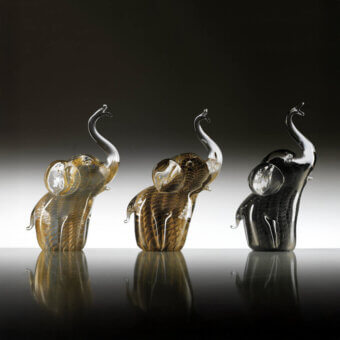 Glass elephant sculptures by Duccio Di Segna—amber, brown, and black—feature plumed trunks and rounded bodies on a gray gradient backdrop.