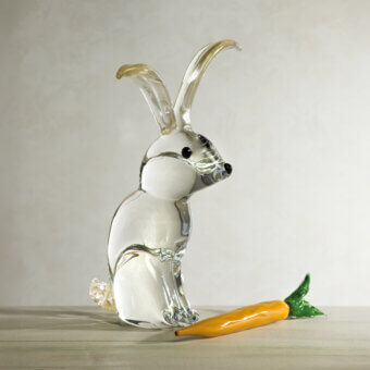 Glass rabbit sculpture with amber ears and black bead eyes, seated beside a glass carrot on a light gray surface.