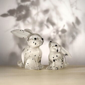 Glass rabbit sculptures, speckled with black, sit on a light wood surface against a blurred grayscale abstract background.