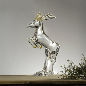 Glass reindeer sculpture, clear with gold accents, rearing pose on wood surface against gray gradient backdrop.