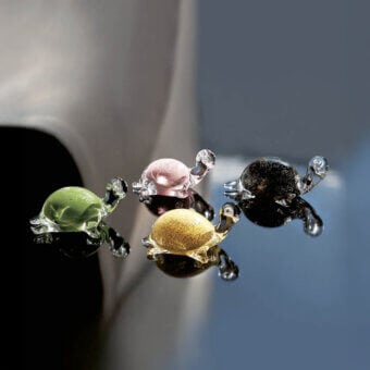 Glass tortoise sculptures in green, pink, black, yellow, and gray hang against a gray and white backdrop, displaying rounded shells and smooth textures.