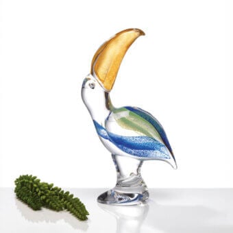 Glass toucan sculpture with blue and green stripes, gold beak, and clear base, displayed on a white reflective surface.