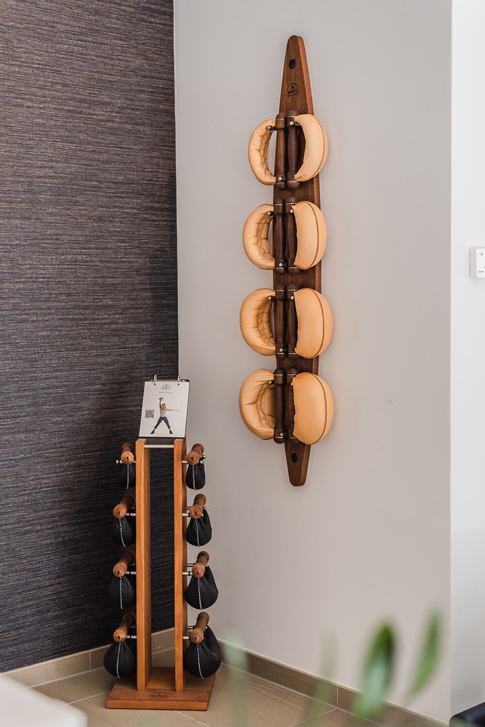 Home gym with walnut wall rack holding tan leather medicine balls, and hardwood dumbbell rack with black weights, against gray and white walls.