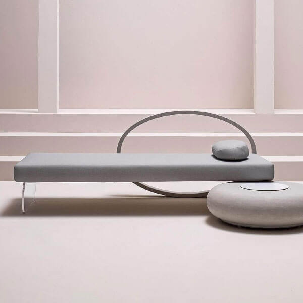 Gray upholstered lounger with clear acrylic legs, set against pale pink paneled walls, includes gray cushion and pouf.