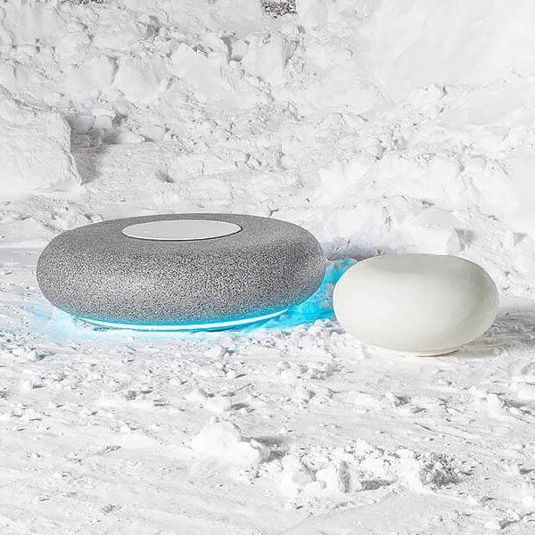 Smart speakers, dark gray and white, circular design with blue light, sit in a snow-covered outdoor scene with natural daylight.