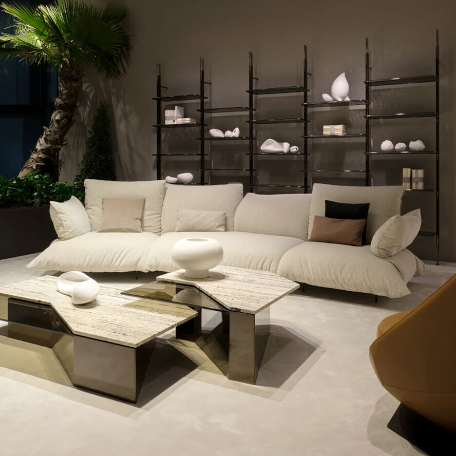 Modular sofa, beige linen, oversized cushions, paired with marble-top coffee tables, shelving unit, palm tree, and armchair in studio setting.