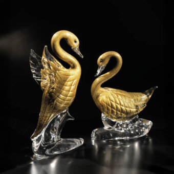 Glass swan sculptures by Duccio Di Segna, gold-toned textured bodies, clear bases, spotlight illumination, fractured wing detail.