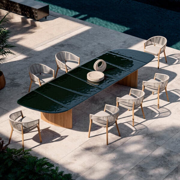 Oval dining table with dark green glass top and woven light gray chairs on a gray tiled patio near a teal pool.