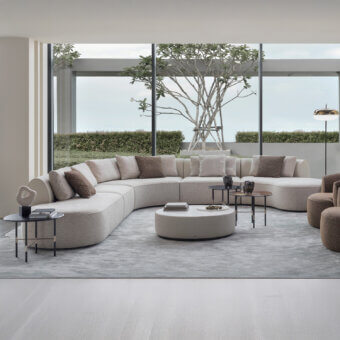 Beige curved sectional sofa with plush cushions, white coffee table, glass side tables, brass floor lamp, gray walls, and green view.