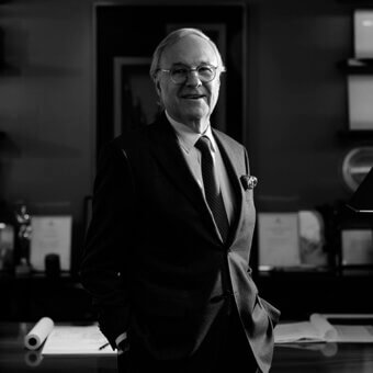 Pierre Yves Rochon portrait, silver hair, dark suit, floral lapel pin, standing in office with desk, drawings, artwork, soft lighting.