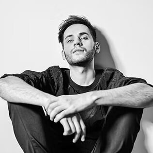 Black and white portrait of Alexey Danilin seated, wearing dark textured clothing, looking upwards with a slight smile against a white background.