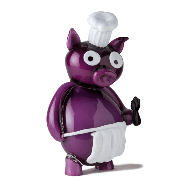 Purple Murano glass pig figurine with chef’s hat and apron, featuring rounded form, white eyes, and raised front leg.