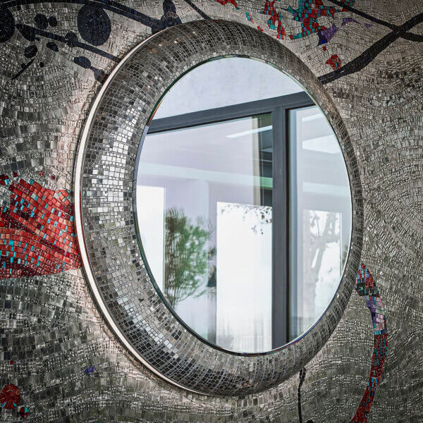 Sicis Fluxus round mirror, silver mosaic tiles, circular frame with red, blue, and black accents, reflecting interior space.