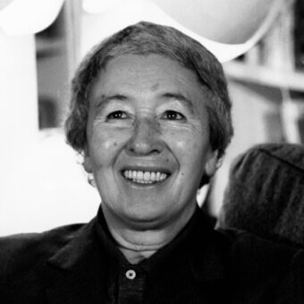 Gae Aulenti portrait, black and white photograph, Italian architect smiling warmly wearing a dark blazer and button-down shirt, blurred interior background.