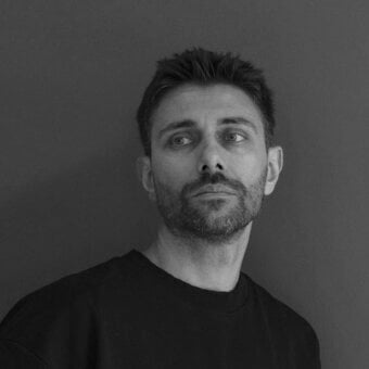 Man portrait, grayscale, short dark hair, black sweatshirt, neutral expression, light gray eyes, solid gray background.