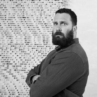 Man with beard, brown shirt, against dense white text backdrop, evenly lit, creating contrast between figure and informational pattern.