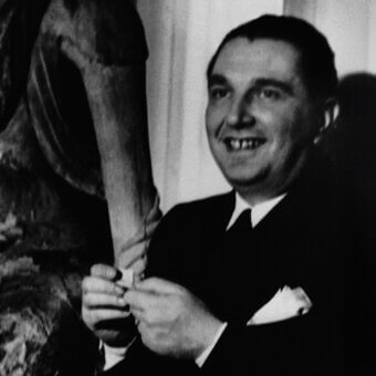 Pietro Chiesa portrait, man in dark suit, standing before textured abstract sculpture with curved arm, soft lighting, neutral background.