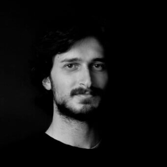Black and white portrait of Simone Di Liddo with dark wavy hair, mustache, and stubble, wearing a dark crew-neck shirt against a black background.