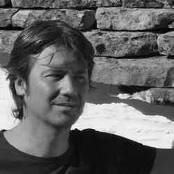 Man portrait against light stone wall, wearing black t-shirt, looking right with a wink, natural lighting, candid composition.