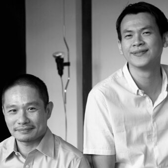 Black and white portrait of Voon Wong & Benson Saw, both wearing light shirts, against a minimalist interior with a suspended microphone.