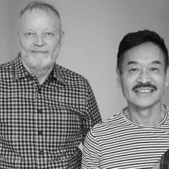 Black and white portrait of two designers, Yabu Pushelberg, with checkered and striped shirts against a gray backdrop.