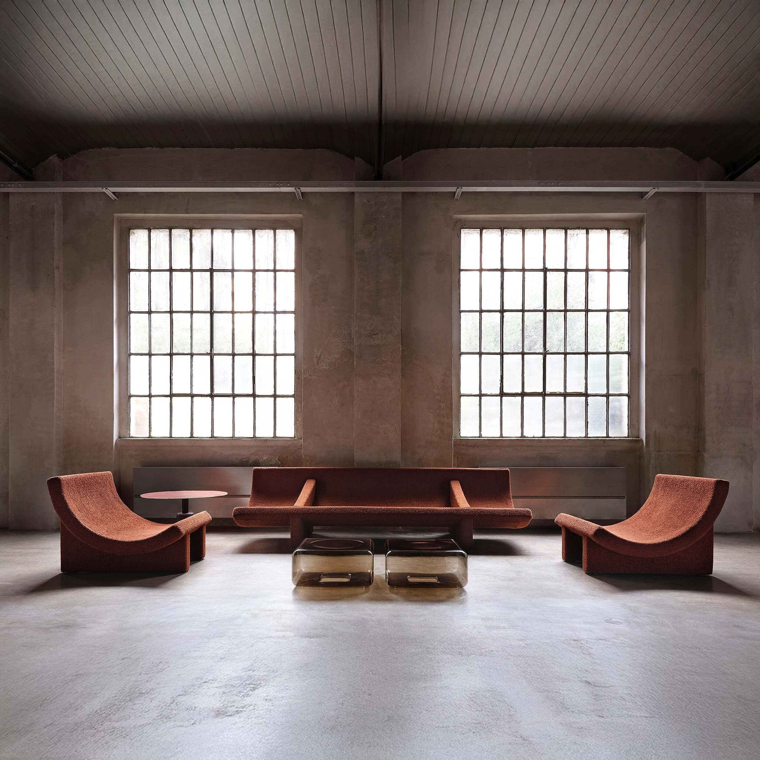 Living room with reddish-brown sofa, wingback chairs, glass coffee tables, and concrete walls, featuring exposed metal beams and barred windows.