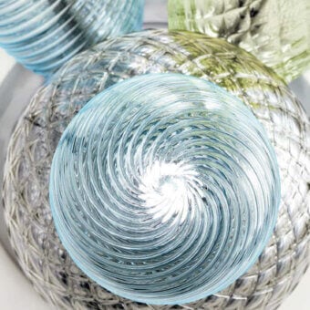 Glass orbs, light blue with spiral pattern, silver woven texture, pale green, and ribbed designs on reflective gray surface.