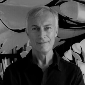 Marco Lucchi portrait with abstract black and white artwork backdrop, stacked papers, soft studio lighting, gray hair, button-down shirt.