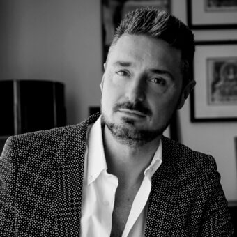 Black and white portrait of Massimiliano Raggi in a patterned black blazer and white shirt, with framed art in background.