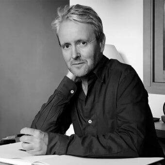 Ronan Bouroullec portrait, black and white photograph, seated man with light blonde hair at a white desk with pen and notebook.