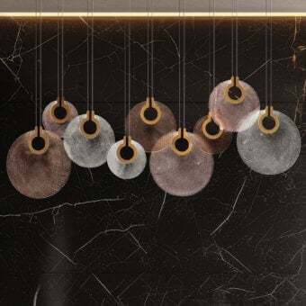 Corallo suspension lamps, perforated metal, gradient gold-silver shades hang before dark gray veined marble, emitting warm diffused light.