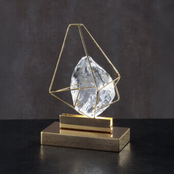 Crystal table lamp, brushed gold metal cage, truncated pyramid shape, clear faceted crystal, dark gray background, reflective black surface.