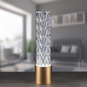 Cassoni table lamp, clear faceted shade, brushed brass base, illuminated against blurred gray interior with sofa and geometric art.