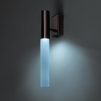 Wall lamp, bronze finish, square mount with ribbed glass shade emitting cool blue light in studio setting.