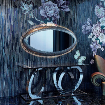Sicis Fluxus mirror, gray mosaic frame, oval shape, mounted on black marble console with silver metal base in a blue textured room.