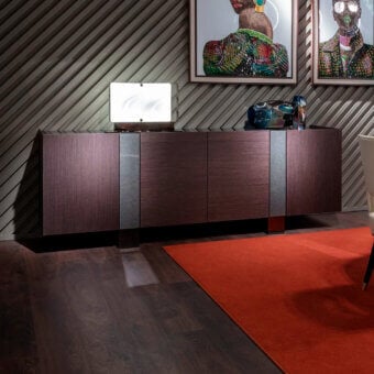 Dark wood sideboard with vertical paneling and striped accents, red rug, textured wall, and white cylindrical lamp.