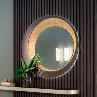 Sicis Harper circular mirror with brushed gold frame, integrated lights, and shelf displaying books and a glass vase with greenery.