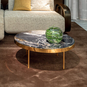 Sicis orbital coffee table, marbled surface with gold veins, brass metal frame, tapered legs, glass globe with foliage, beige sofa visible.