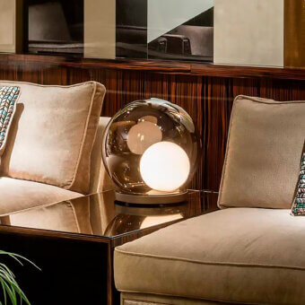 Sicis Rigel table lamp with smoked glass shade and illuminated spheres on dark glossy table, flanked by beige armchairs.