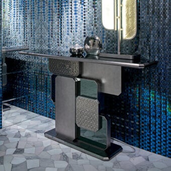 Marble console table, dark gray metal finish, chevron base with mirrored accents, blue mosaic tile backdrop, glass sphere on surface.