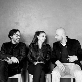 Black and white portrait of three people seated on wood stools against a textured neutral wall, wearing dark jackets and sweaters.