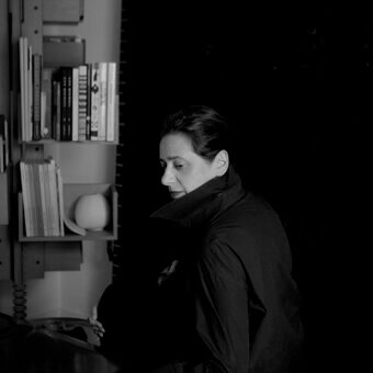 India Mahdavi portrait, grayscale, seated at desk with books on metal shelving, wearing dark collared shirt, soft directional lighting.