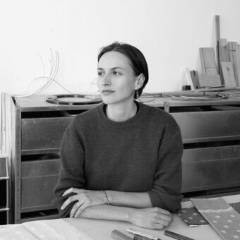 Black and white portrait of Julie Richoz seated at a table with tools, a patterned fabric, and a metal cabinet backdrop.