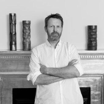 Martino Gamper portrait with textured cylindrical sculptures on a weathered mantel, soft lighting, and neutral wall backdrop.