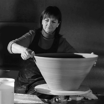 Pottery studio scene with woman shaping large ceramic bowl on wheel, wearing green shirt and apron, soft diffused lighting.