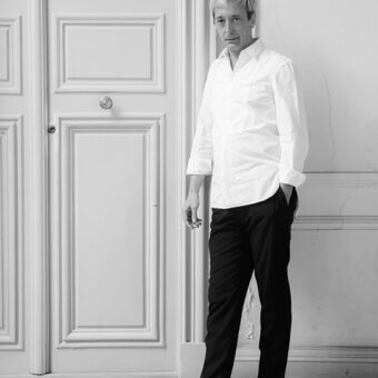Black and white portrait of Sam Baron in white shirt, standing before paneled door with rectangular sections and tiled floor.