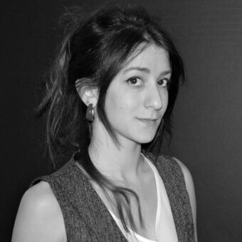Black and white portrait of Francesca Borelli, gray vest, voluminous hair, direct gaze, dark gray background, smooth skin tone, even lighting.